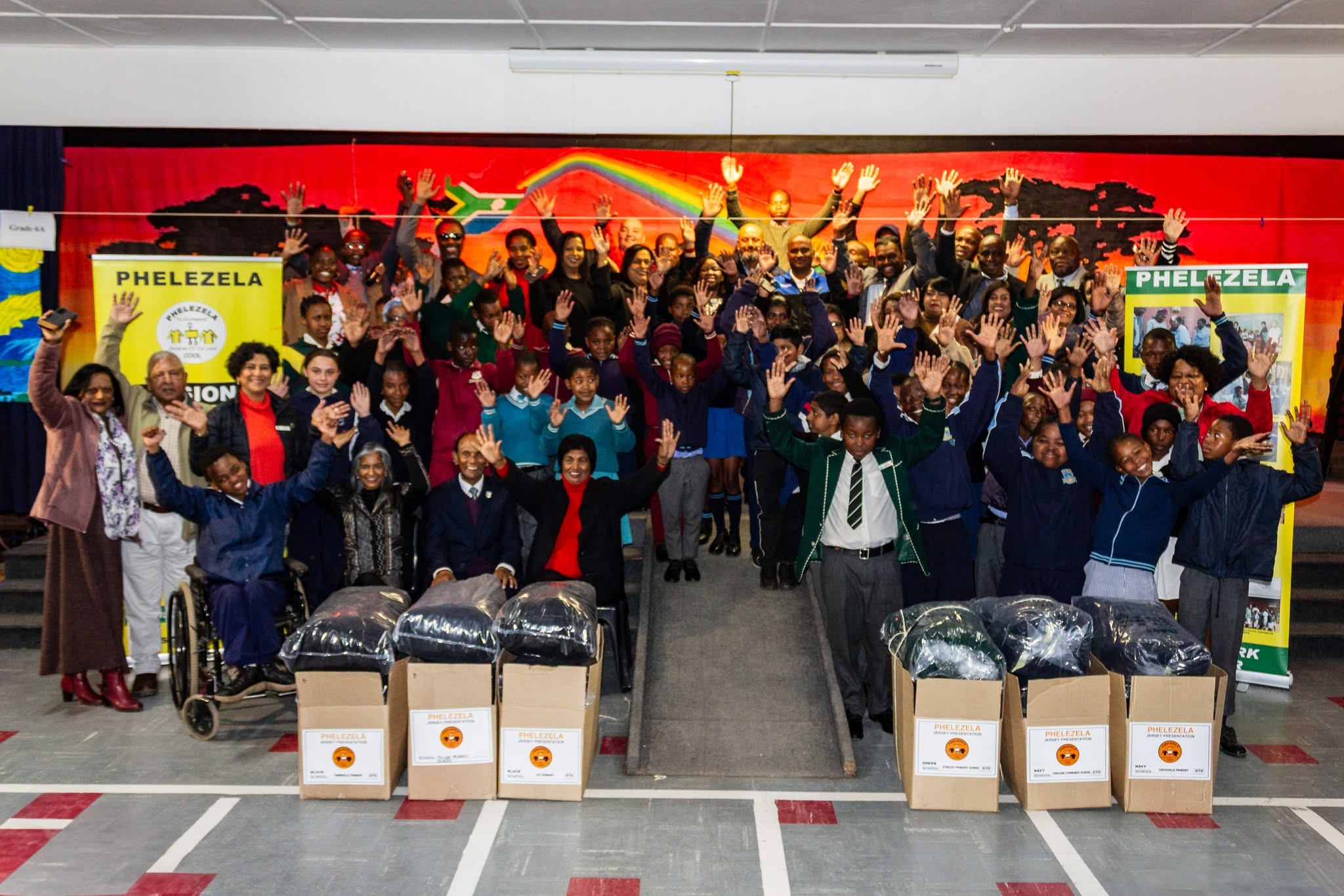 School children, teachers and Phelezela committee members celebrating with raised hands behind boxes of new jerseys at the 2025 Jersey Presentation